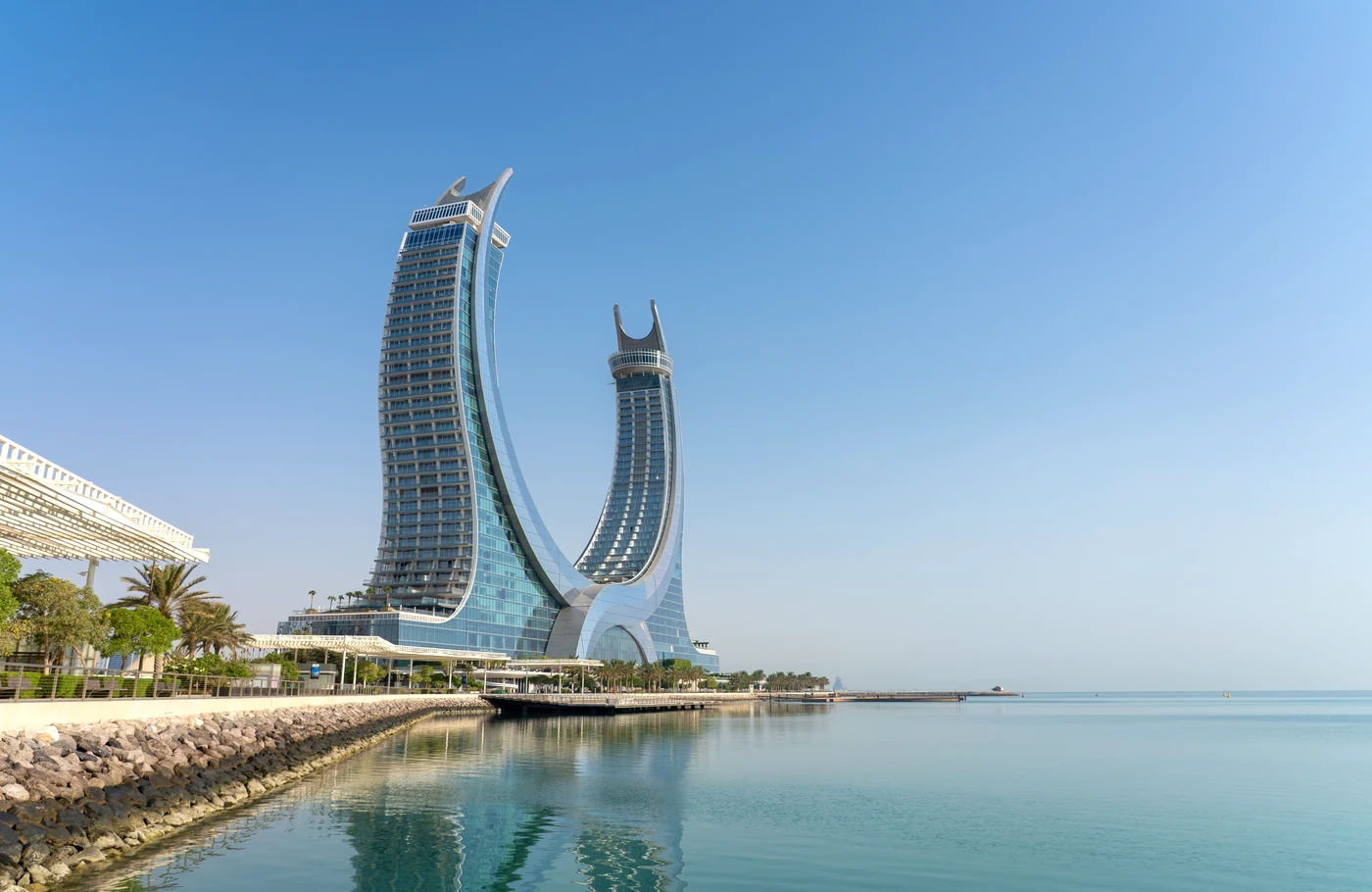 Katara Towers beside the Lusail waterfront, reflected in calm water under a clear blue sky